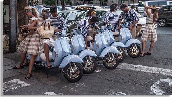 foto com diversos jovens vestidos como na década de 50, com motocicletas azuis do tipo vezpa enfileiradas