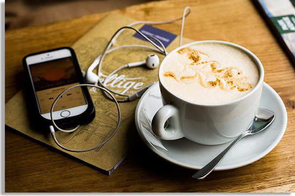 mesa de madeira com bule, colher de chá e caneca preenchida de café com leite. Ao lado, um celular com fones de ouvido